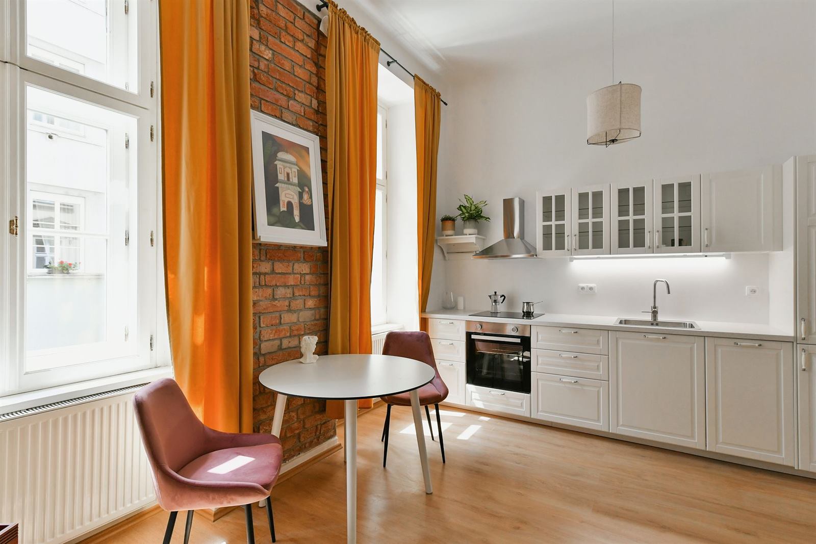 Dining table by exposed brick wall and amber curtains, with fully equipped white kitchen